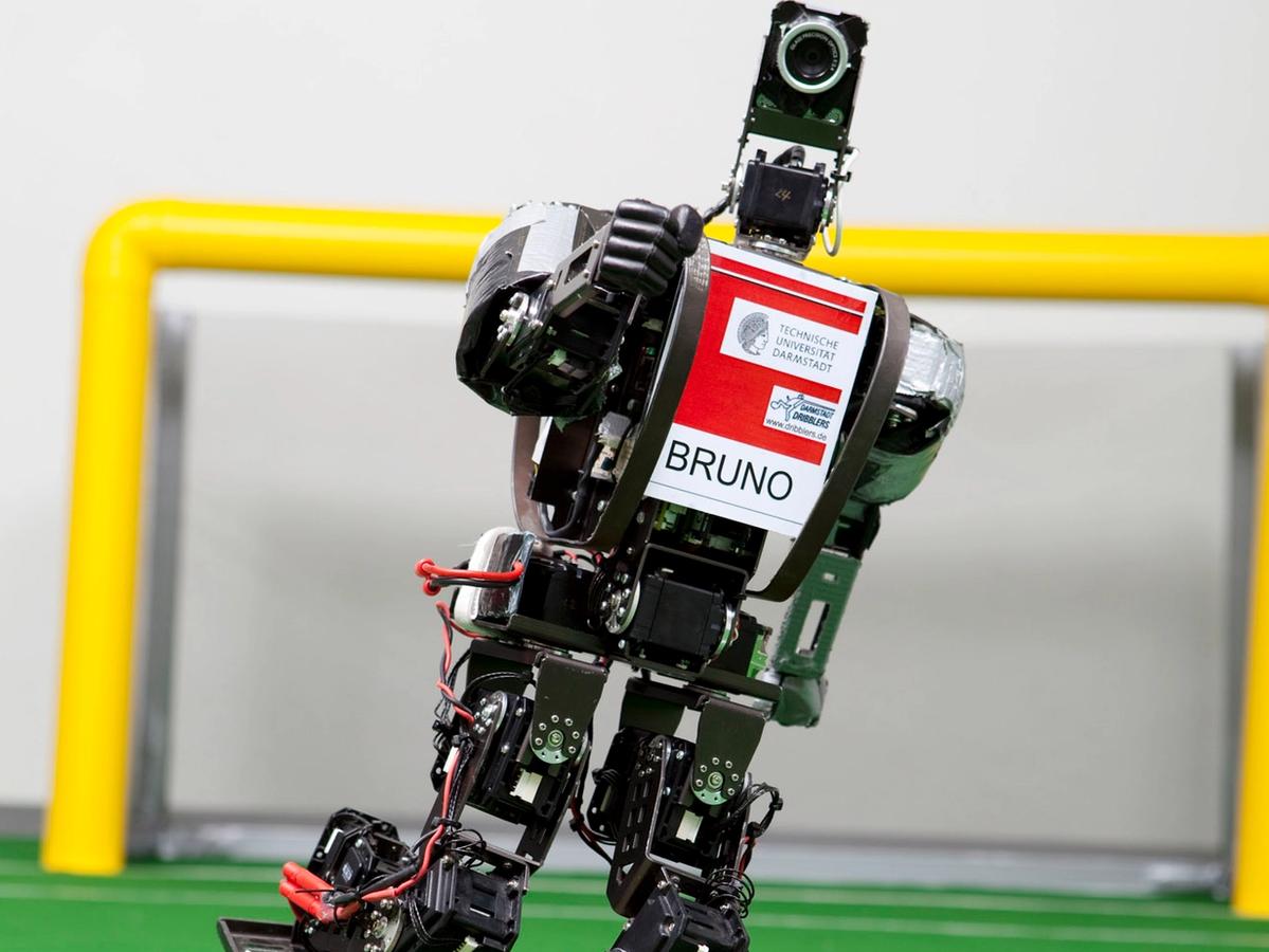 A black two-legged robot labelled Bruno kicks a orange ball on a miniature soccer field.