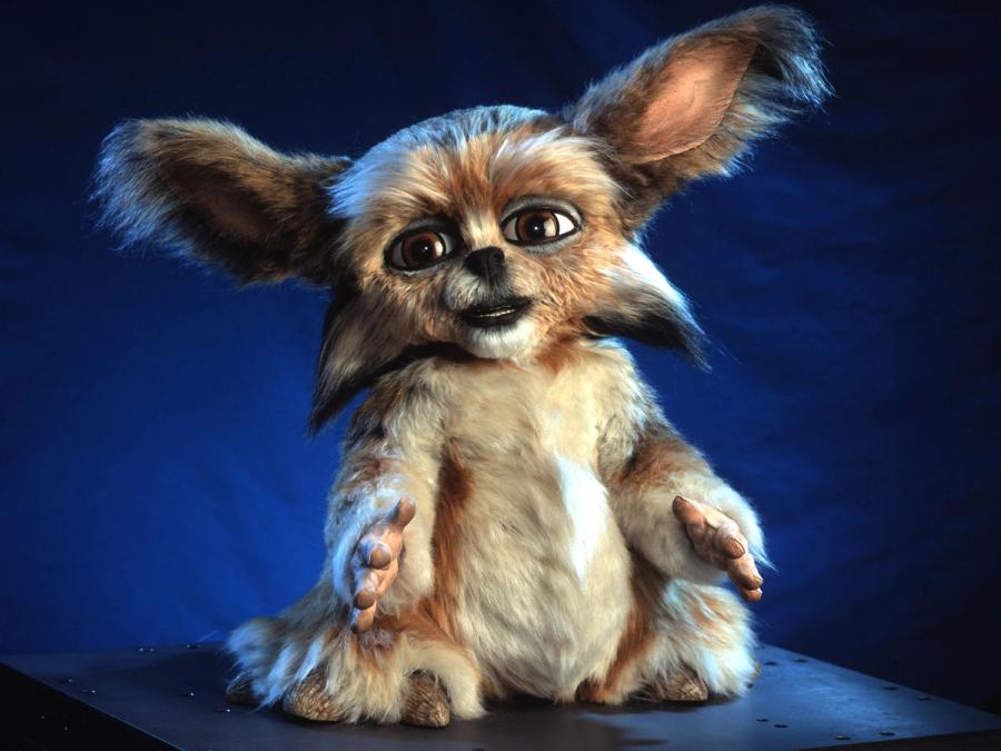 A seated furry robot with big brown eyes, a dog-like face with a black nose and teeth, and protruding ears. It's body is covered in white and brown fur and it has hands with four fingers.