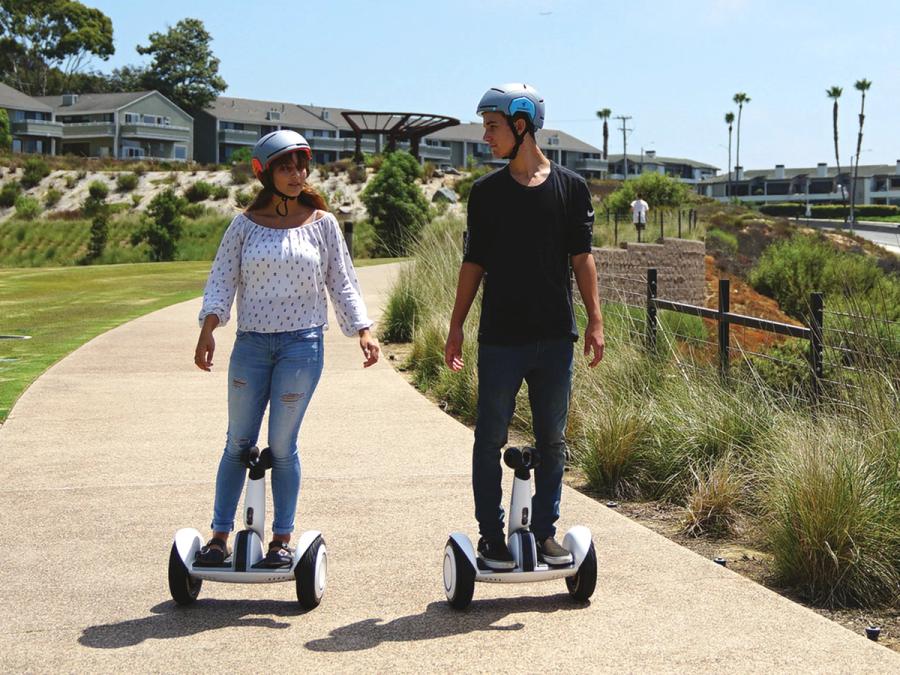 Two people in helmets ride mini Segways which include the base on two wheels and a vertical piece balanced between the rider's legs. 
