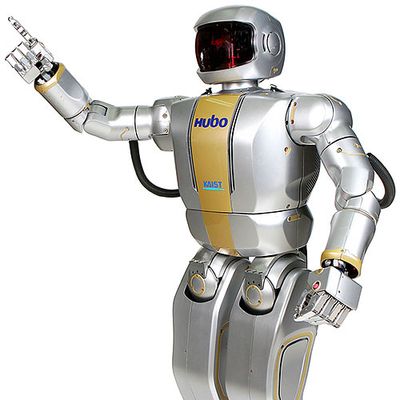 A sleek silver robot with head like an astronauts helmet holds one arm up and points one of its fingers.