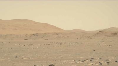 Ingenuity helicopter seen very small on the left bottom of the frame flying over the rocky rusty surface of Mars.