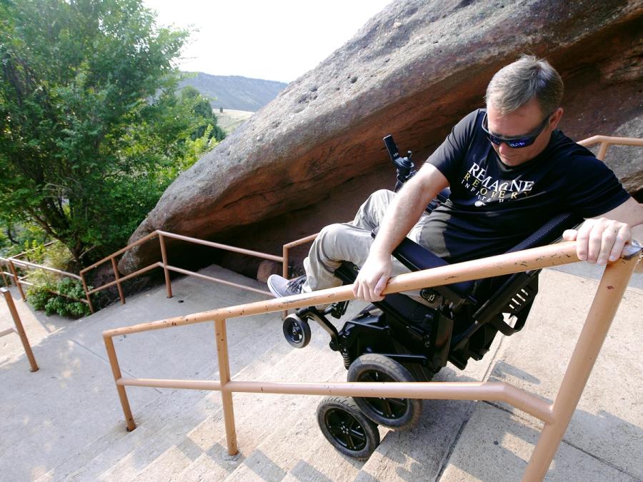 A man sits in a robotic wheelchair that is elevated on its back wheels, and is on the steps of a long outdoor set of stairs. His hands are braced on a guardrail to move himself.