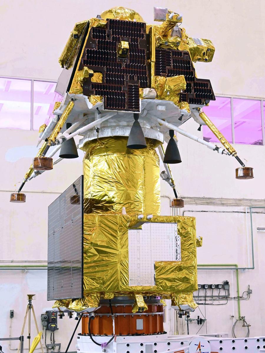 A large space lander sits atop other parts of a craft, with gold foil and solar panels, in a white lab environment.