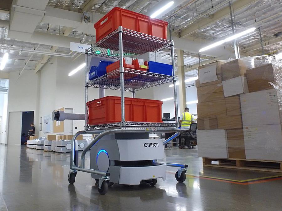 A white mobile robot carries a three tiered shelf with red and blue bins in a factory setting.