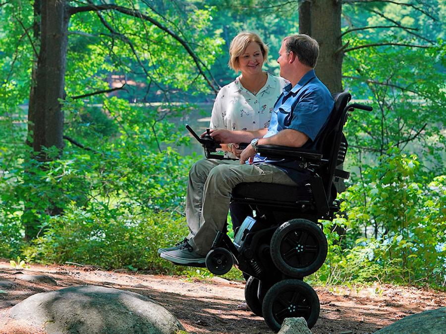 A smiling woman faces a man who sits in a robotic wheelchair that is elevated on two of its four main wheels. They are on an outdoor trail, surrounded by green leafy trees.