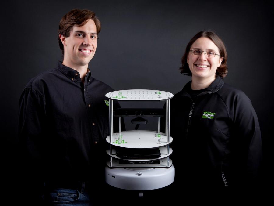 A man and a woman in black shirts stand holding a multi-tiered circular white robot.