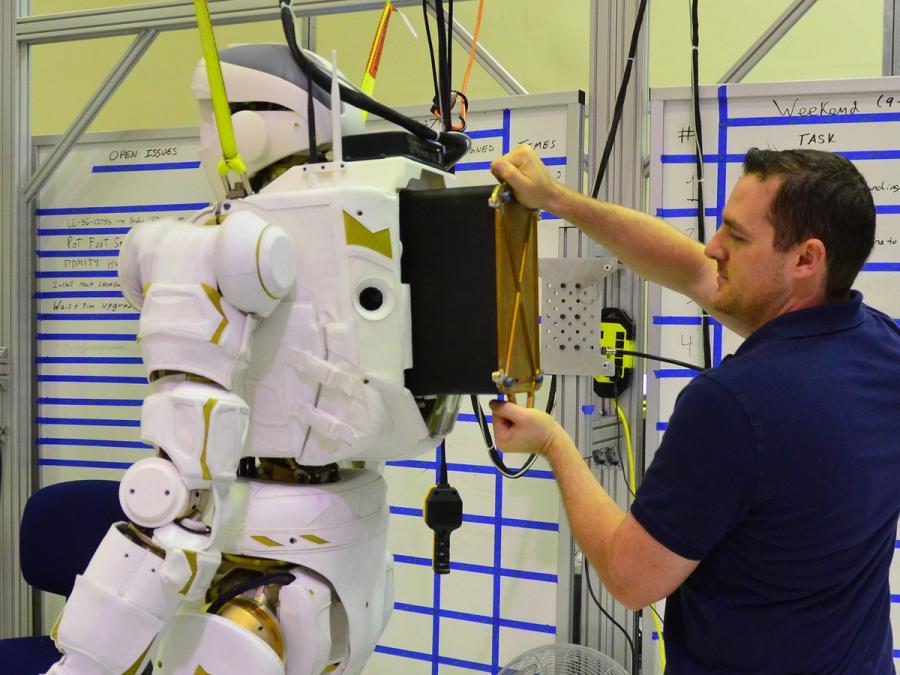A man adjusts a large black and gold backpack on Valkyrie, who is held up by a harness in a lab.