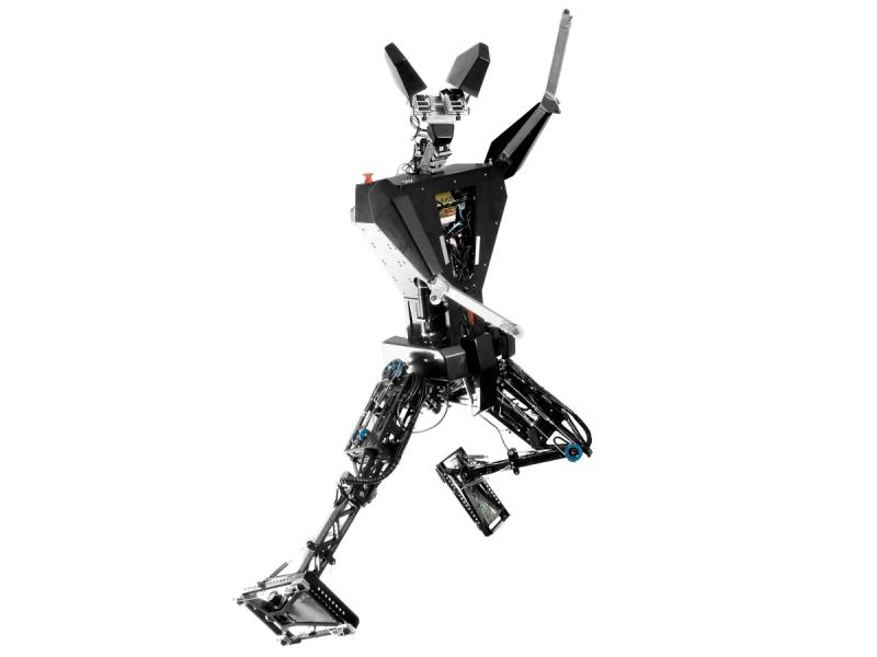A humanoid with an electronics filled torso the shape of an upside down triangle, a silver head with sensors and two large black ears that stick out of the top of its head. It has two basic silver arms, one of which is raised. It's legs are posed dynamically such that it is balanced on one leg which is thrust out to the left, while leaning to the right.