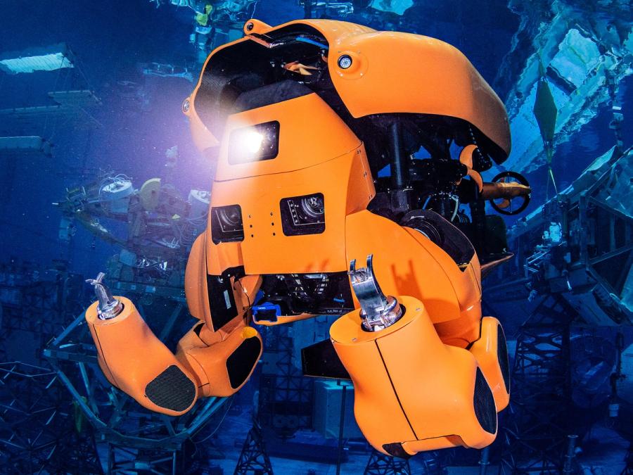 An orange half humanoid submarine robot with gripper hands shines a light underwater.