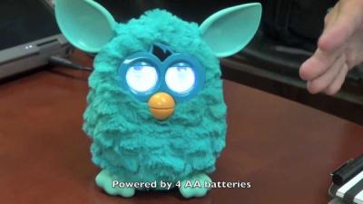 A green Furby stands on a wooden desk with its eyes lighting up.