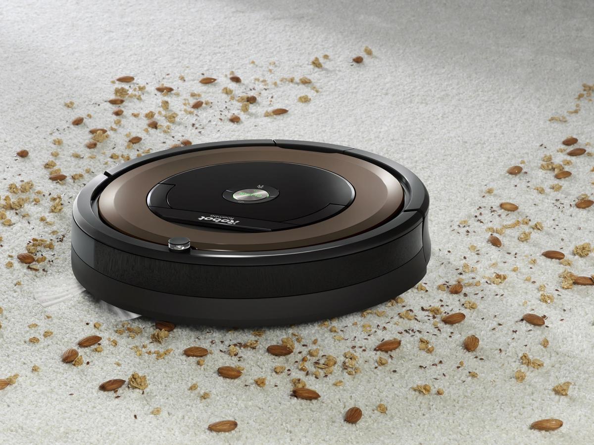 A squat robotic vacuum cleans a carpet covered in food debris.