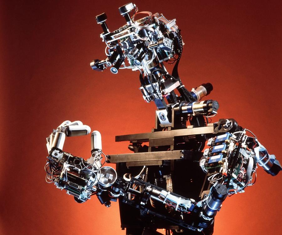 A human shaped robot with exposed electronics looks at it's fingers, which are pushed together to form an O.