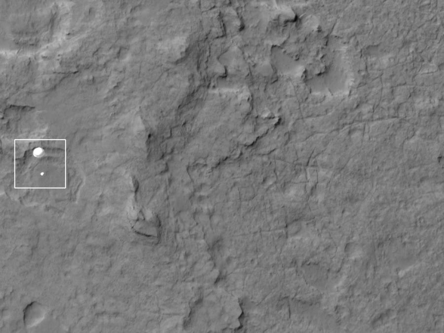 A black and white image of the surface of Mars with a white square highlighting a parachute approaching the ground.