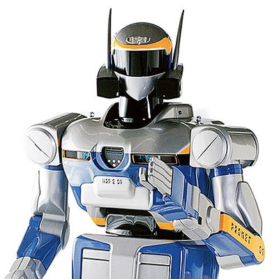 A bipedal humanoid robot with a blue, silver, black and yellow exterior and the appearance of wearing a helmet is posed with its arms and gripper fingers ready to act.