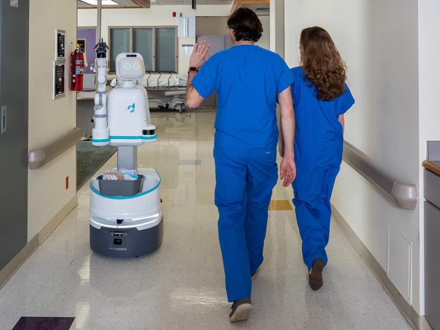 A white robot with glowing eyes, a gripper arm, and a mobile base, waves at two hospital employees.