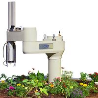 A white robotic arm with a vertical appendage sticks out from a flower filled circular garden in a platform.