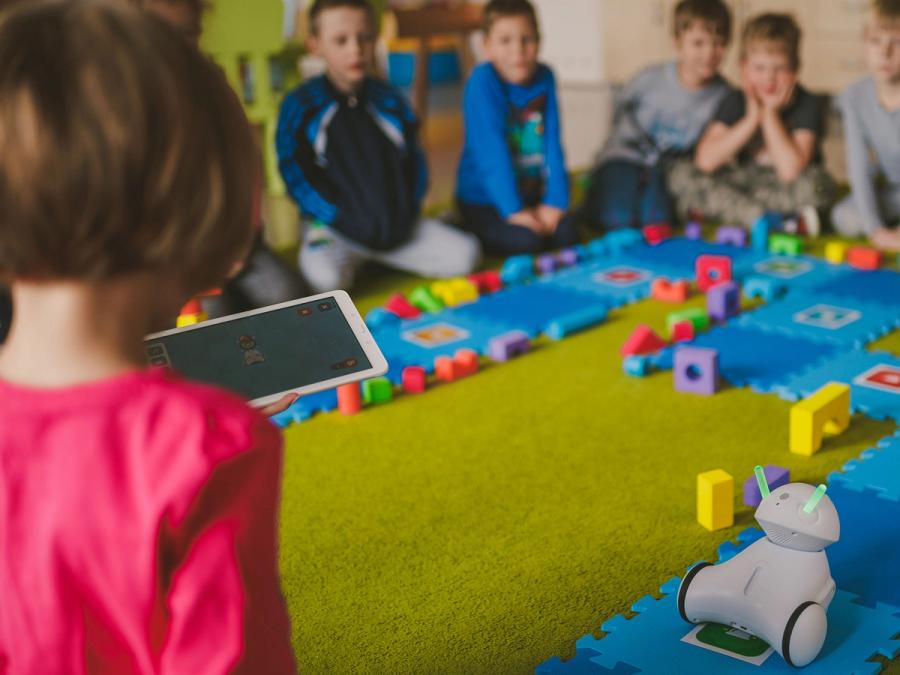 A group of children look at a Photon robot as it moves through a simple obstacle course. A girl with a tablet is in the foreground.