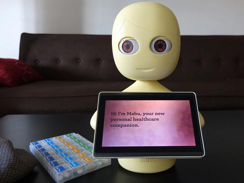 A simple yellow robot with a tabletop base supporting an egg shaped head which has big brown eyes. It holds a tablet which has text on it. A container of pills is next to it.