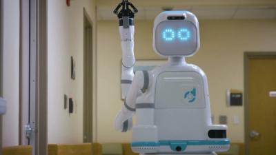 A white robot with LED eyes stands at a hospital hallway with its gripper hand up in the air.