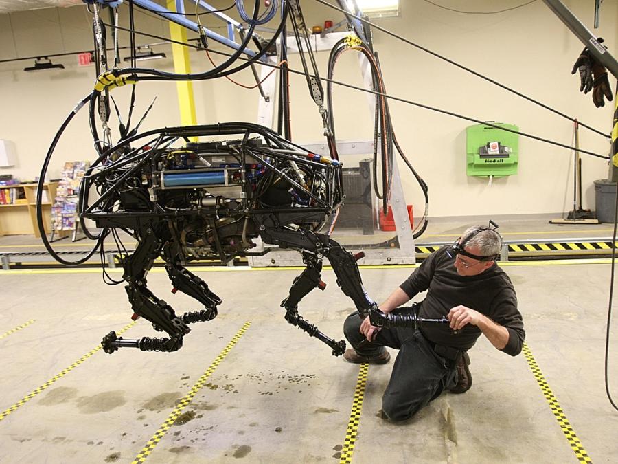 A quadruped robot is lifted on a harness while a man uses a tool on it's leg.