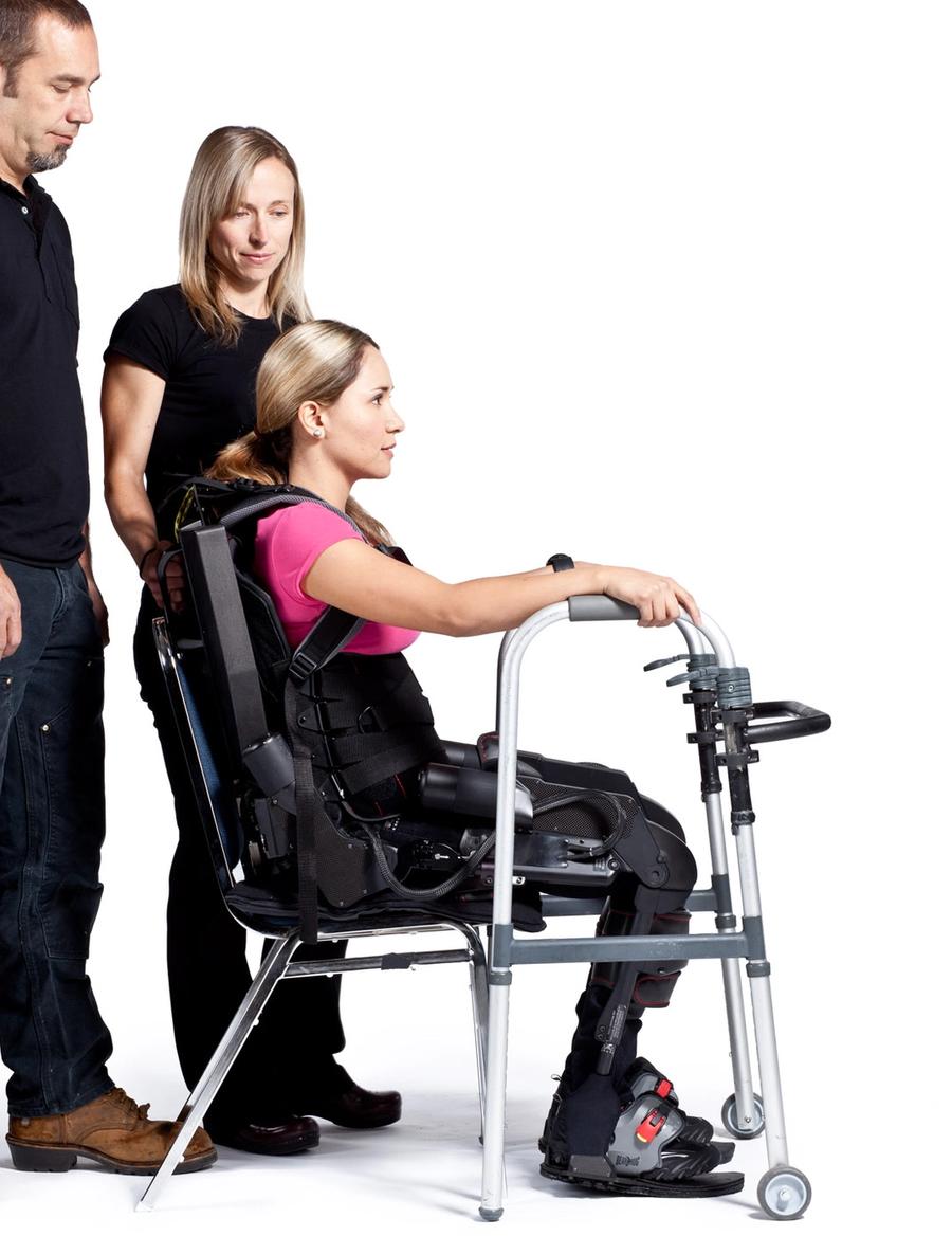 The subject is seated while wearing the exoskeleton, and has her hands on a walker.