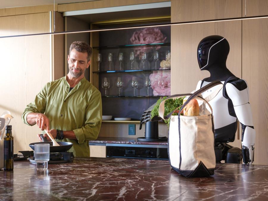 Tesla's Optimus, a silver and black bipedal humanoid robot, stands at a kitchen counter next to a man who is cooking.