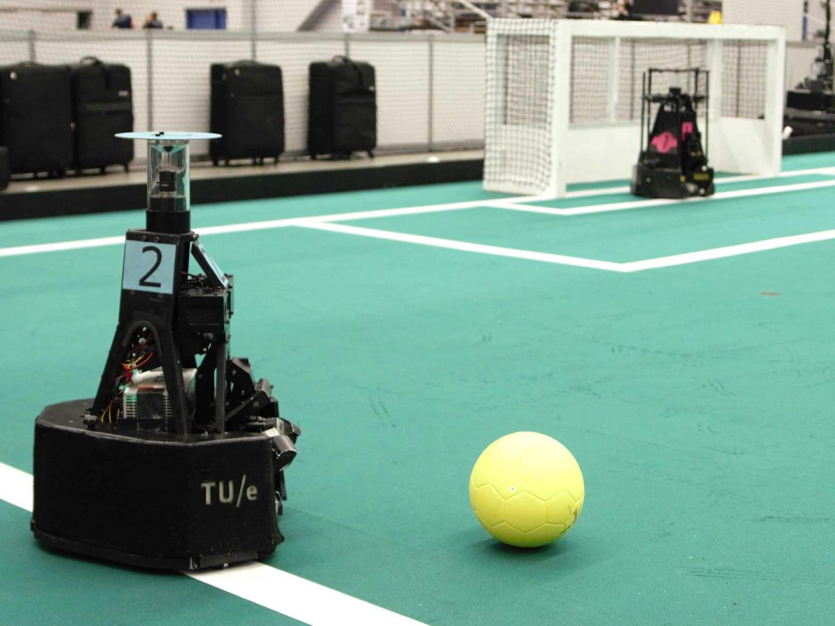 Two black mobile robots play soccer on a green field. One is nearing the soccer ball while another is in the goal box.