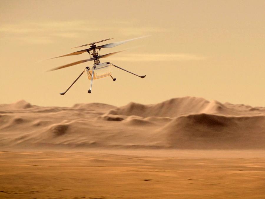 A small in-flight helicopter on Mars, with a square silver body the size of a tissue box, a pair of carbon fiber rotors, a rectangular solar panel on top, and four spindly legs on the bottom.