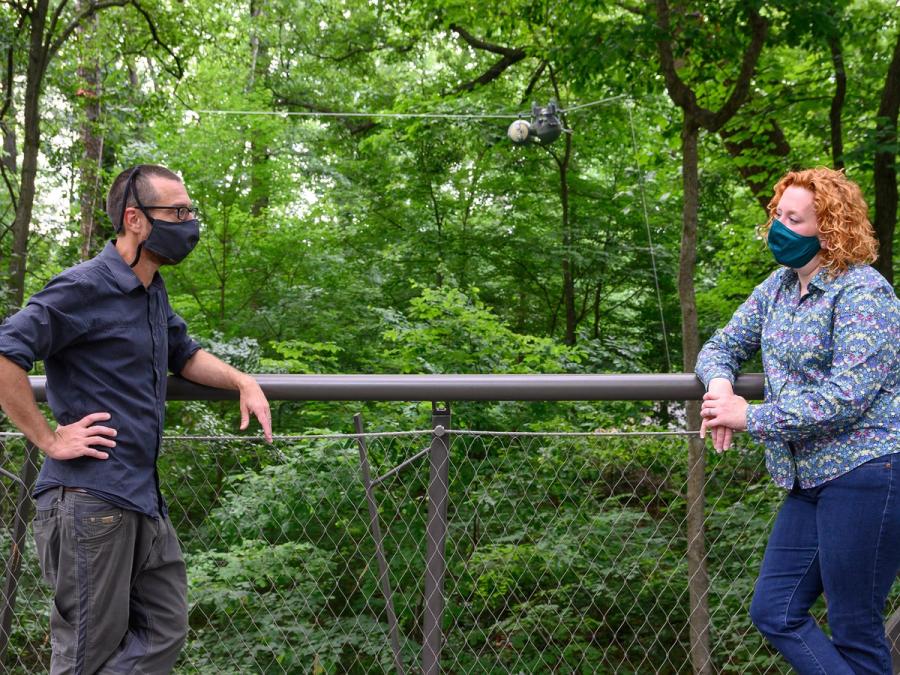 Two people wearing masks lean on a fence outdoors. In the distance behind them, a sloth robot hangs from a wire.