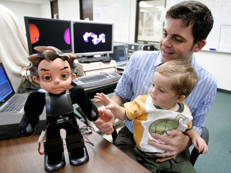 A man with a young toddler on his lap interact with a small humanoid robot sitting on top of a desk.