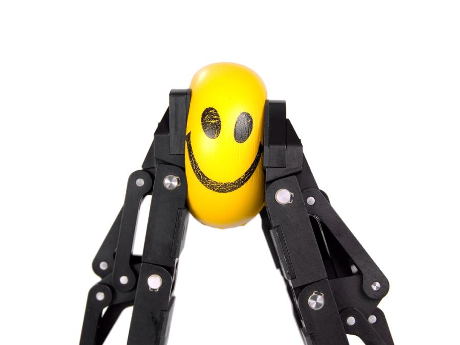 A pair of black robotic grippers smush a yellow smiley face ball.