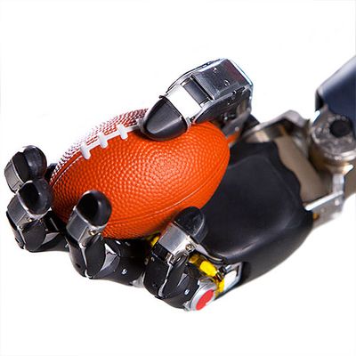 A black robotic hand with five jointed fingers mimicking a human hand holds an orange miniature football.