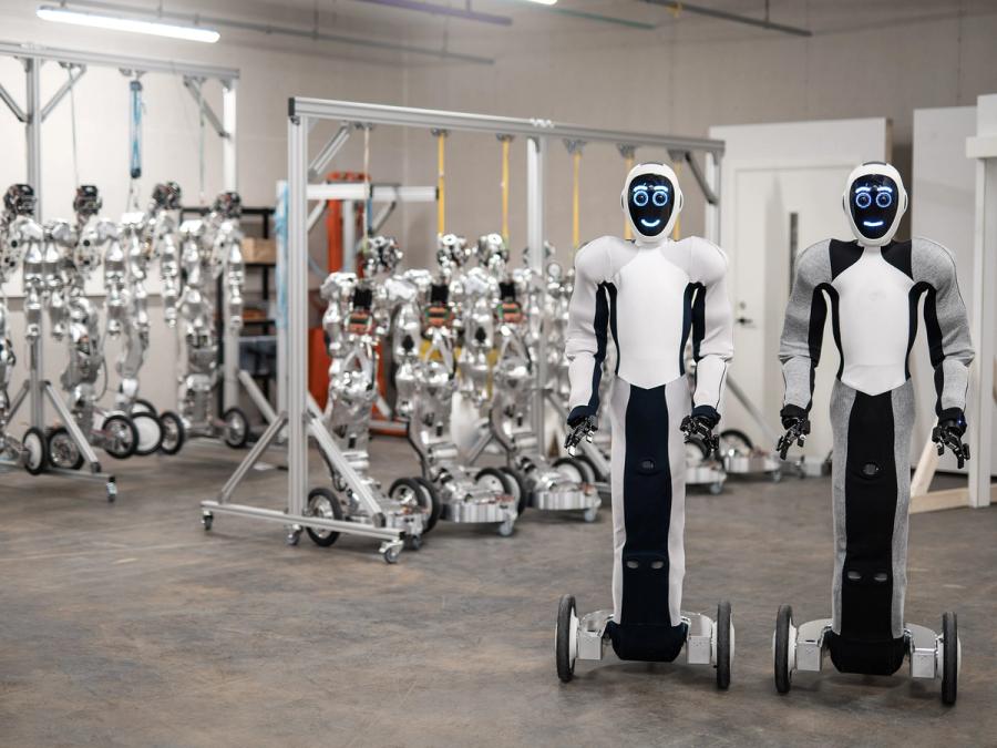 Two white and black humanoid robots on wheeled bases, with gripper arms, and display faces showing round eyes and smiling mouths, stand in front of rows of humanoid body structures.