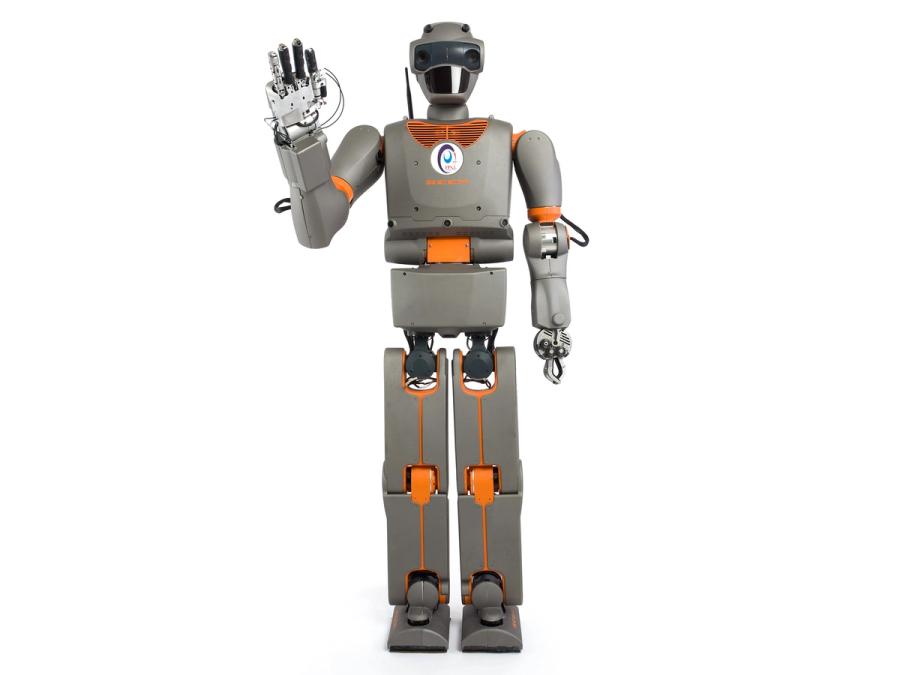 A grey and orange humanoid waves at the camera. It has a cartoonish head with eyes and a plate over its mouth area. The hand that waves has four fingers, and the other hand has a gripper end effector.