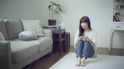 A woman with long brown hair, white shirt, jean, and barefoot sits on a living room floor staring to Qoobo, a furry pillow-shaped robot with a tail and no head, which is on a couch next to her.
