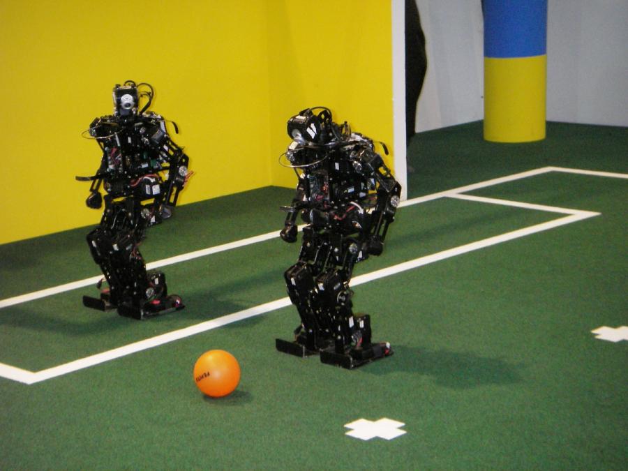 Two black robots on miniature soccer field look at an orange ball.