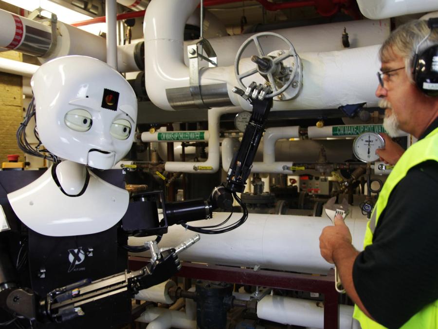 The robot is in a room with many white pipes. It gestures at a man wearing safety headphones and vest.