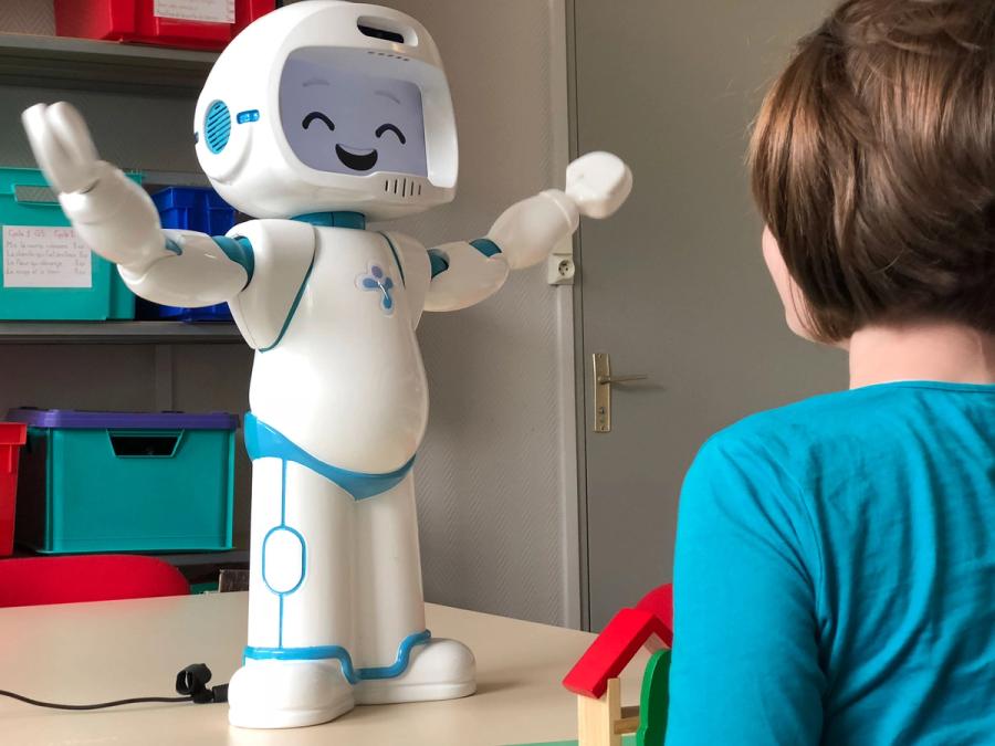 The robot stands on a tabletop with its arms extended outward and a laughing expression on its face. A child is looking at it.