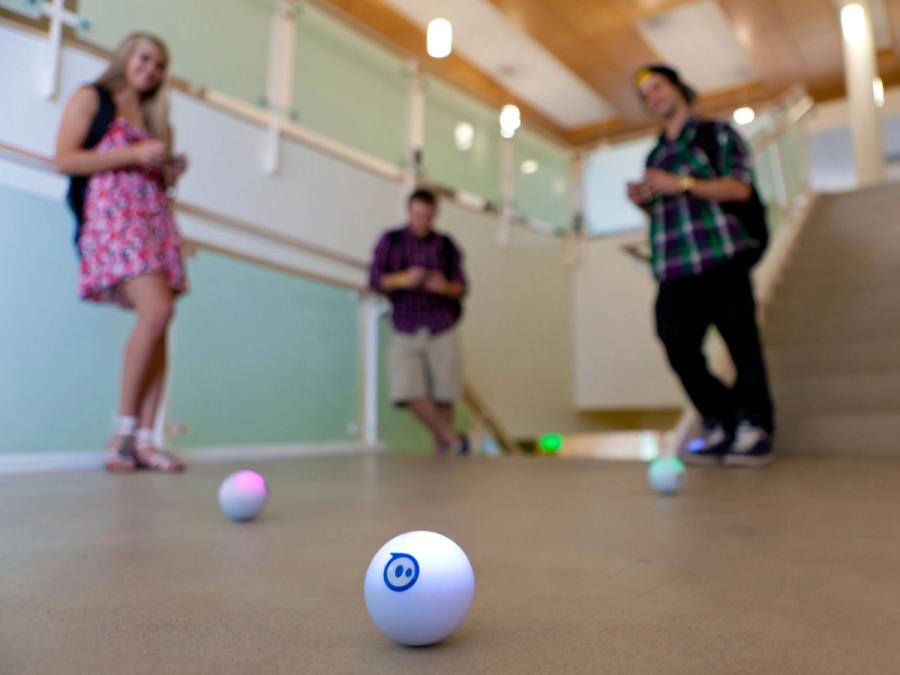 Three teenagers in the background control three white Sphero ball robots from devices.