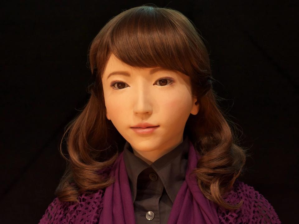 A highly realistic humanoid female robot with golden skin, almond shaped eyes, and shoulder length brown hair wears a black button up shirt and purple sweater.