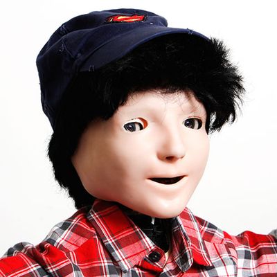 Close-up on the face of a child-size humanoid with peachy flesh fitted over camera eyes and open mouth. It wears a black wig, blue cap and plaid shirt.
