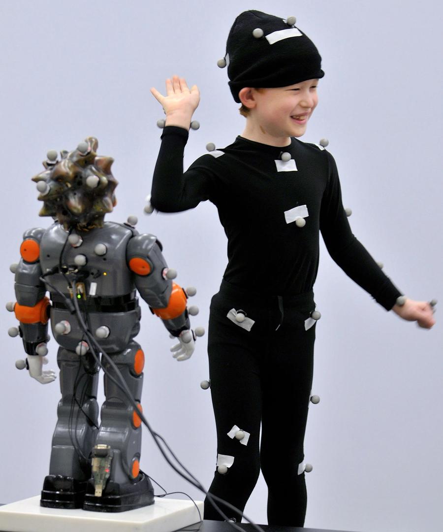A smiling child waves while wearing a full body black suit and hat next to small humanoid robot on a platform.