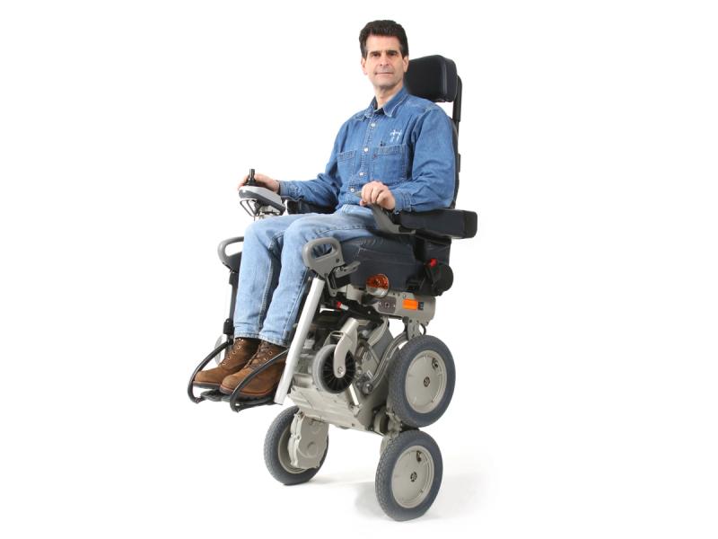A man clothed in denim shirt and jeans sits in a robotic wheelchair that is elevated on two of its four main wheels.