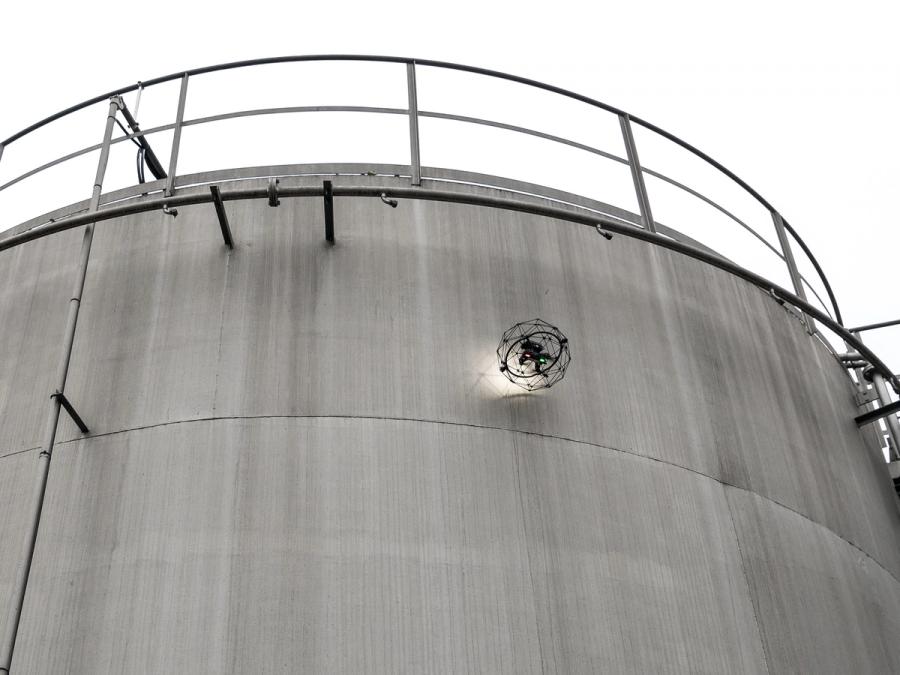 A drone in a circular protective cage flies up to the top of an industrial structure to do inspection.