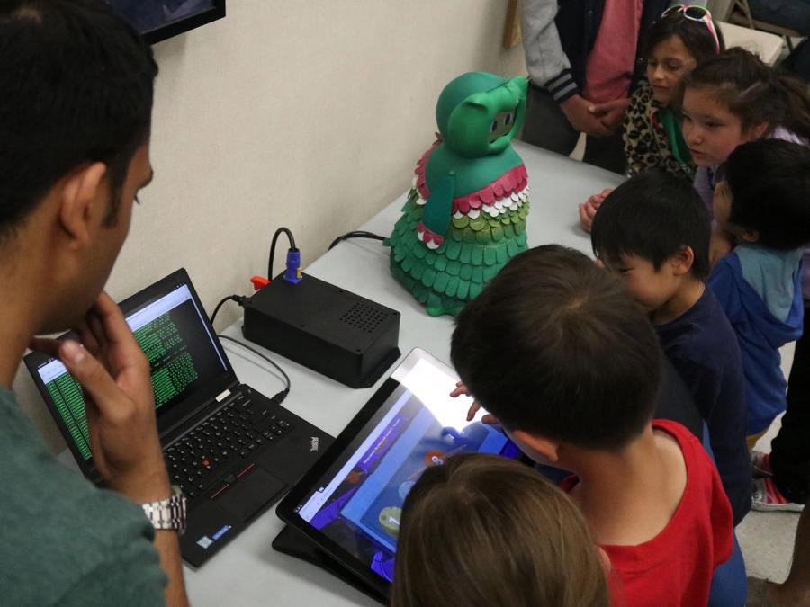 The robot sits on a table surrounded by children looking at computers that will control it.