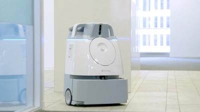 A gray and white plastic robotic vacuum slightly bigger than an upright vacuum drives itself at an office.