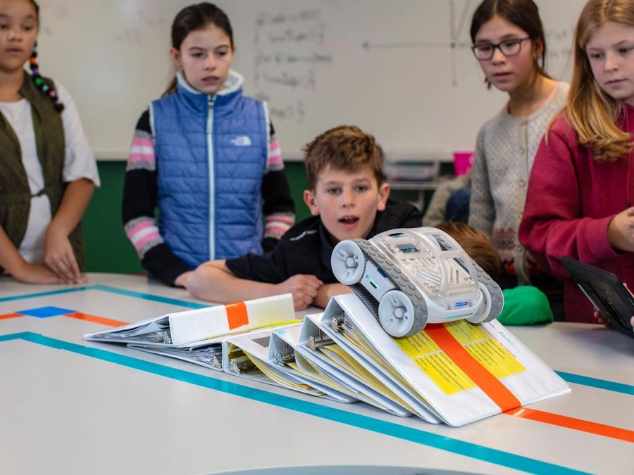 A robotic vehicle rides down some loosely stacked binders while children look on in amazement.