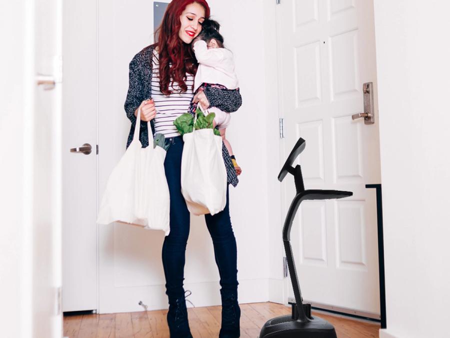 A woman holding a toddler and carrying groceries looks at Temi, a telepresence robot that is about half of her height.