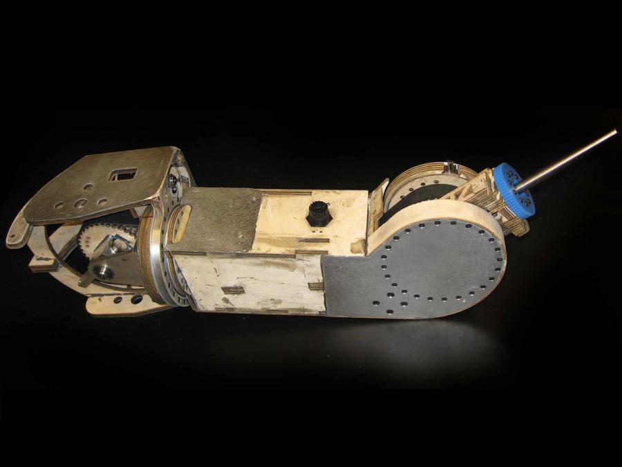 The robot arm is seen on on a black background. It is mostly made of plywood, with some aluminum pieces.