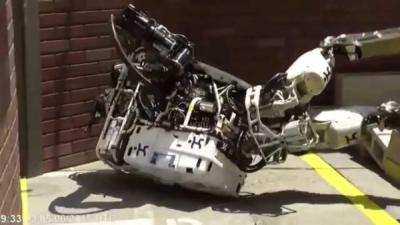 Atlas, a large humanoid robot with metal frame and exposed cables and actuators, falls on its back on a concrete floor at a robotics competition arena.